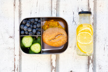 Bottle Of Lemonade And Lunch Box With Cucumber Slices, Blueberries And Bun With Lentil Paste