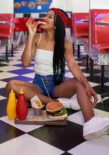 Young Woman With Braided Hairstyle Sitting On The Floor With Her Hamburger Plate And Eating A Chip