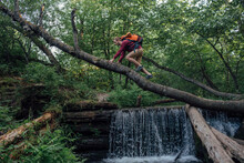 Young Hiker With Backpack Crossing Water On Tree Trunk In The Forest