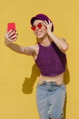 Wall Mural - Non-binary person smiling while taking selfies with a mobile phone outdoors against a yellow wall.