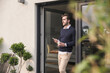 © UWE_UMSTAETTER/Westend61 - Young man leaning in door of his house, holding cup of coffee, using smartphone