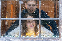 Smiling Siblings Looking At Snowfall Through Window While Sitting At Home