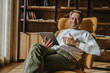 © Mareen Fischinger/Westend61 - Mature man holding digital tablet while sitting on chair against shelf in living room