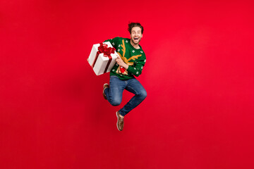 Poster - Full size photo of good mood positive guy jumping hold new year present wear ugly sweater isolated on red color background