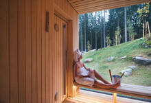 Relaxing Woman Wrapped White Towel Sitting On The Wooden Bench In Hot Finnish Sauna With A Huge Wide Window With Green Forest View And Enjoying Pleasant Healthy Body Care Temperature Treatment.