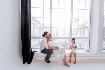 Wall Mural - Happy family is having fun while sitting on the windowsill by the panoramic window. Woman pregnant