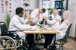 © sofiko14 - Team of multi ethnic doctors clapping to male colleagues after speech during conference. Happy medical specialists with one in wheelchair sitting at boardroom and coworking.