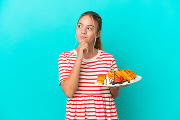 Wall Mural - Little caucasian girl holding waffles isolated on blue background and looking up