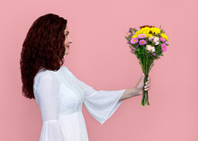 Woman Offering Flower Bouquet With Soft Smile Isolated On Pink Background. Side View Portrait Of Beautiful Woman Giving Flowers To Someone In Front Of Her. She Has Wavy Red Brown Hair.