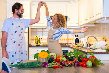 Happy adult couple dance and have fun together in the kitchen at home while preparing healthy vegetables on the table. Overjoyed woman and man in love prepare lunch and enjoy relationship