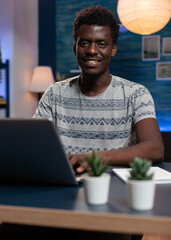Poster - Portrait of smiling african american entrepreneur man browsing management information on laptop computer. Young freelancer working remote at online project in living room late at night