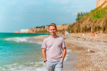 Wall Mural - A young blond man walks on the beach near the azure ocean or sea. The concept of a happy summer vacation