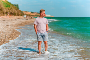 Wall Mural - A young blond man walks on the beach near the azure ocean or sea. The concept of a happy summer vacation