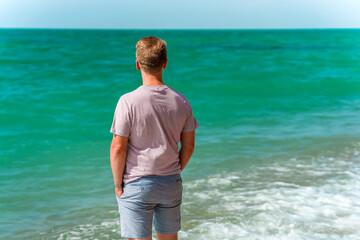 Wall Mural - A young blond man walks on the beach near the azure ocean or sea. The concept of a happy summer vacation