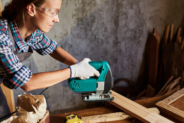 Female Carpenter sawing wood with an electric jigsaw. Woman in apron doing some carpentry work in his workshop or garage, using electric jigsaw.