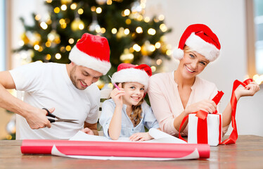 Wall Mural - winter holidays, leisure and family concept - happy smiling mother, father and daughter in santa hats packing gift box and cutting wrapping paper at home over christmas tree lights on background