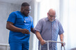 © Acronym - Caregiver is teaching old man to walk with walker. Professional nurse and patient in a nursing home. Assistance, rehabilitation and health.