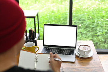 Poster - Behind view of a creative man taking notes while sitting at the wooden working desk surrounded by a computer laptop, coffee cup and various equipment.