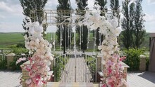 Square Wedding Arch Is Decorated With Artificial Flowers And Shiny Hanging Beads. White Path Leads To Arch On Sides Of Which Are Wooden Chairs. Natural Landscape In Background