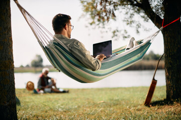 below view of man works on laptop while relaxing in hammock by lake.