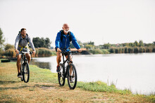 Happy Senior Athlete And His Son Ride Bicycles By The Lake In Nature. Copy Space.