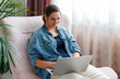 © Beton Studio - Beautiful smiling young woman working on laptop at home interior on comfortable sofa chair near window with plants. Freelancer Modern business woman use laptop for remote work or online shopping