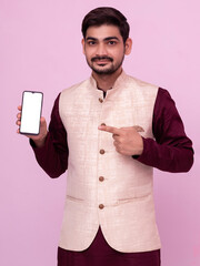 Portrait of  man with beard in traditional wear pointing at cell phone and smiling at camera. Young Indian man in traditional wear and showing smartphone.