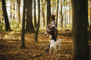 Sticker - Stylish woman in hat and with backpack walking with cute dog in sunny autumn woods. Young female traveler hiking with swiss shepherd white dog in forest. Travel with pet