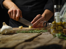 A Chef In A Black Uniform Slices Garlic On A Cutting Board. A Sprig Of Rosemary And Olives Stand On A Wooden Table. Preparation Of Salad, Soup, Sauce, Salad, Pizza, Focaccia. Macro Photography.