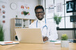 © sofiko14 - African american doctor in eyeglasses and lab coat sitting at desk, typing on laptop and smiling on camera. Concept of technology, medicine and profession.
