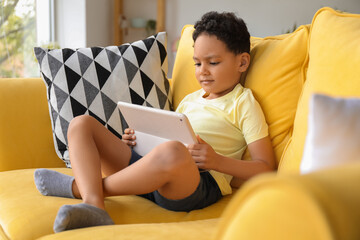 Poster - Little African-American boy with tablet computer watching cartoons on sofa at home