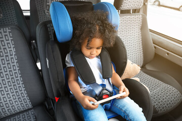 Canvas Print - Little African-American girl with mobile phone watching cartoons in car safety seat