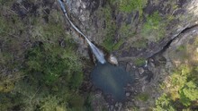 White Waterfall With Foam On Large Rocky Cliff With Moss And Grass Against Small Lake By Brown Stones In Summer Aerial View