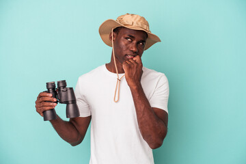 Wall Mural - Young African American man holding binoculars isolated on blue background relaxed thinking about something looking at a copy space.