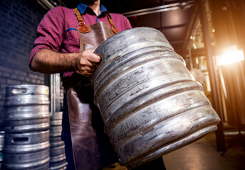 Wall Mural - Young male brewer in leather apron holds barrel with craft beer at modern brewery factory