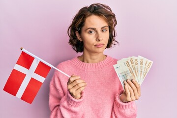 Sticker - Young brunette woman holding norway flag and krone banknotes relaxed with serious expression on face. simple and natural looking at the camera.