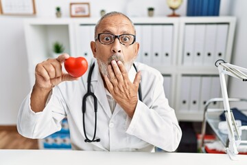 Poster - Mature doctor man holding red heart at the clinic covering mouth with hand, shocked and afraid for mistake. surprised expression