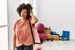 African american woman with afro hair holding yoga mat at pilates room confuse and wondering about question. uncertain with doubt, thinking with hand on head. pensive concept.