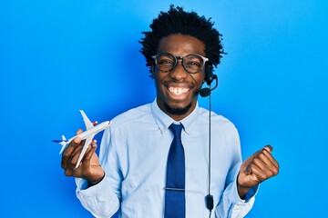 Poster - Young african american travel agent man holding plane screaming proud, celebrating victory and success very excited with raised arm