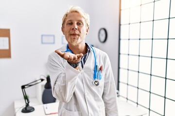 Canvas Print - Young blond man wearing doctor uniform and stethoscope at clinic looking at the camera blowing a kiss with hand on air being lovely and sexy. love expression.