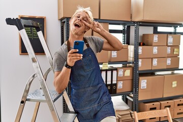 Sticker - Young blond man using smartphone working at storehouse doing peace symbol with fingers over face, smiling cheerful showing victory