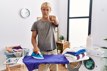 Poster - Young blond man ironing clothes at laundry room pointing with finger to the camera and to you, confident gesture looking serious