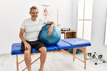 Sticker - Middle age hispanic man at pain recovery clinic holding pilates ball winking looking at the camera with sexy expression, cheerful and happy face.