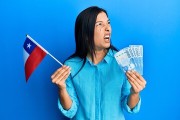 Sticker - Young latin woman holding chile flag and chilean pesos banknotes angry and mad screaming frustrated and furious, shouting with anger. rage and aggressive concept.