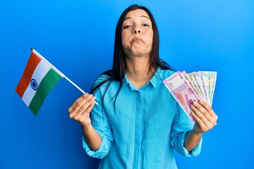 Sticker - Young latin woman holding india flag and rupee banknotes puffing cheeks with funny face. mouth inflated with air, catching air.