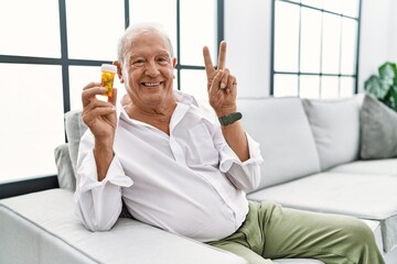 Wall Mural - Senior man holding pills smiling looking to the camera showing fingers doing victory sign. number two.