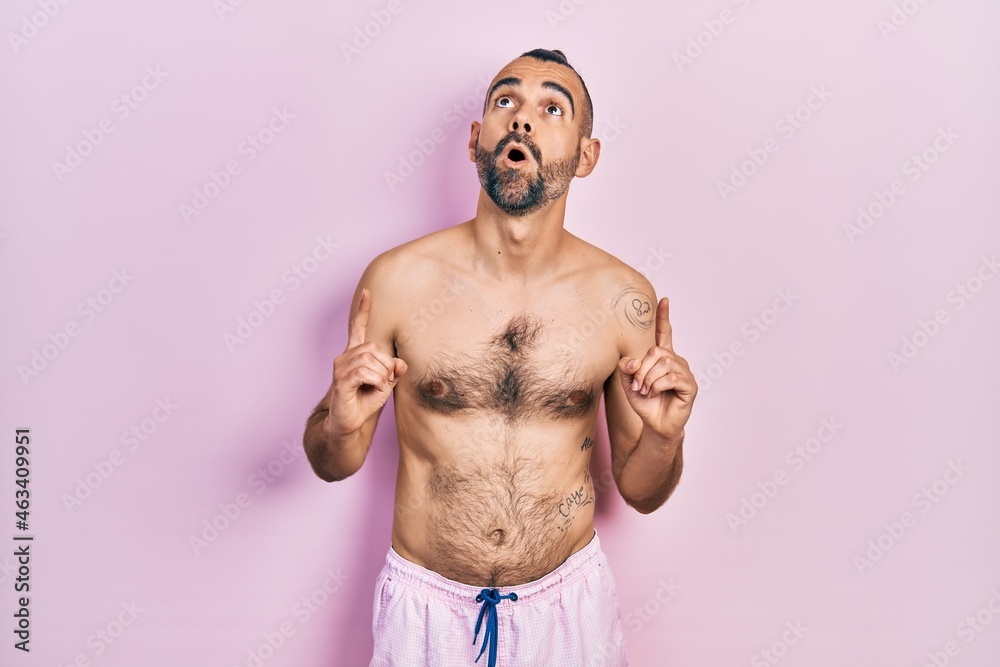 Young hispanic man shirtless wearing swimsuit amazed and surprised looking up and pointing with fingers and raised arms.