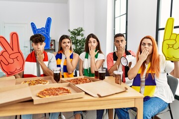 Sticker - Group of young hispanic people eating pizza supporting soccer team at home covering mouth with hand, shocked and afraid for mistake. surprised expression