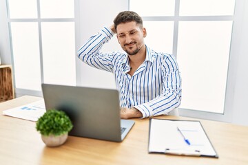 Young handsome man with beard working at the office using computer laptop confuse and wonder about question. uncertain with doubt, thinking with hand on head. pensive concept.