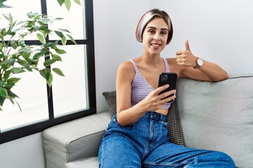 Sticker - Young beautiful woman using smartphone typing message sitting on the sofa doing happy thumbs up gesture with hand. approving expression looking at the camera showing success.